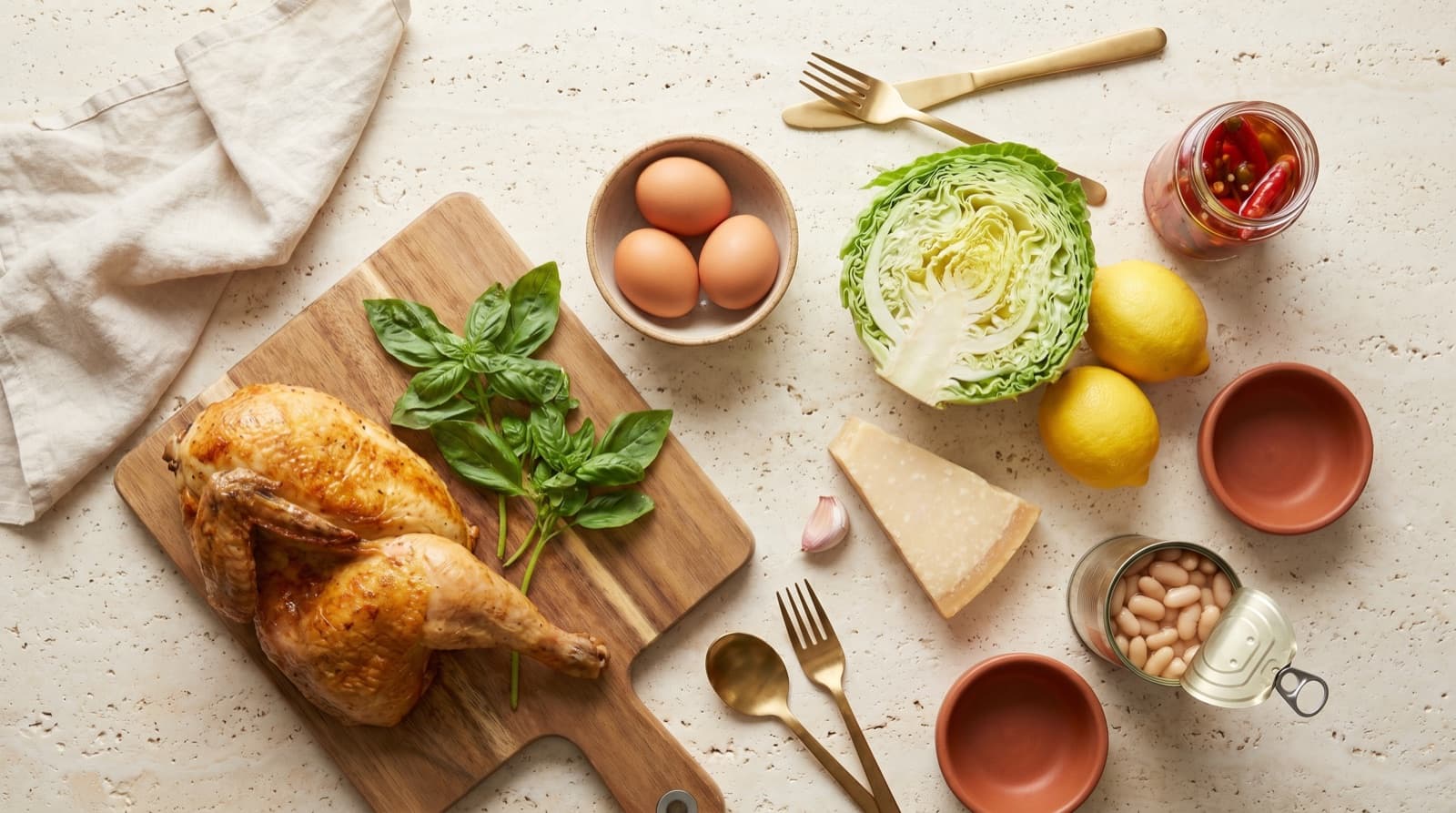 An overhead inventory shot of random fridge-and-pantry ingredients — half a roast chicken on a wooden board, fresh basil, lemons, halved cabbage, eggs, garlic, pickled chillies, beans and parmesan on cream linen